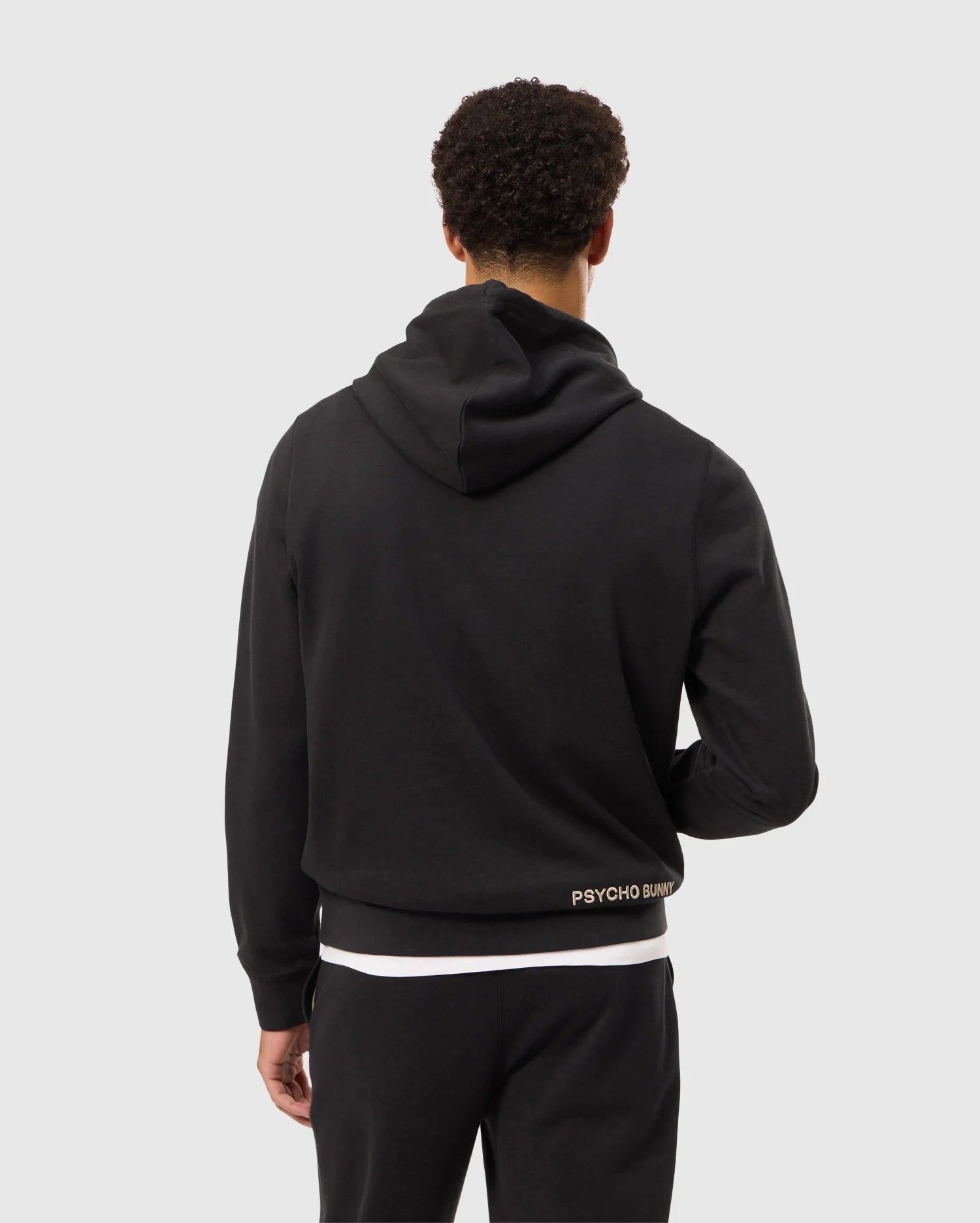 A person with short curly hair is shown from the back, wearing a black hoodie and black pants. The MENS JAMES PREMIUM HOODIE - B6H789D200 from Psycho Bunny features an embroidered bunny logo near the lower back that reads "Psycho Bunny." The background is plain light gray.