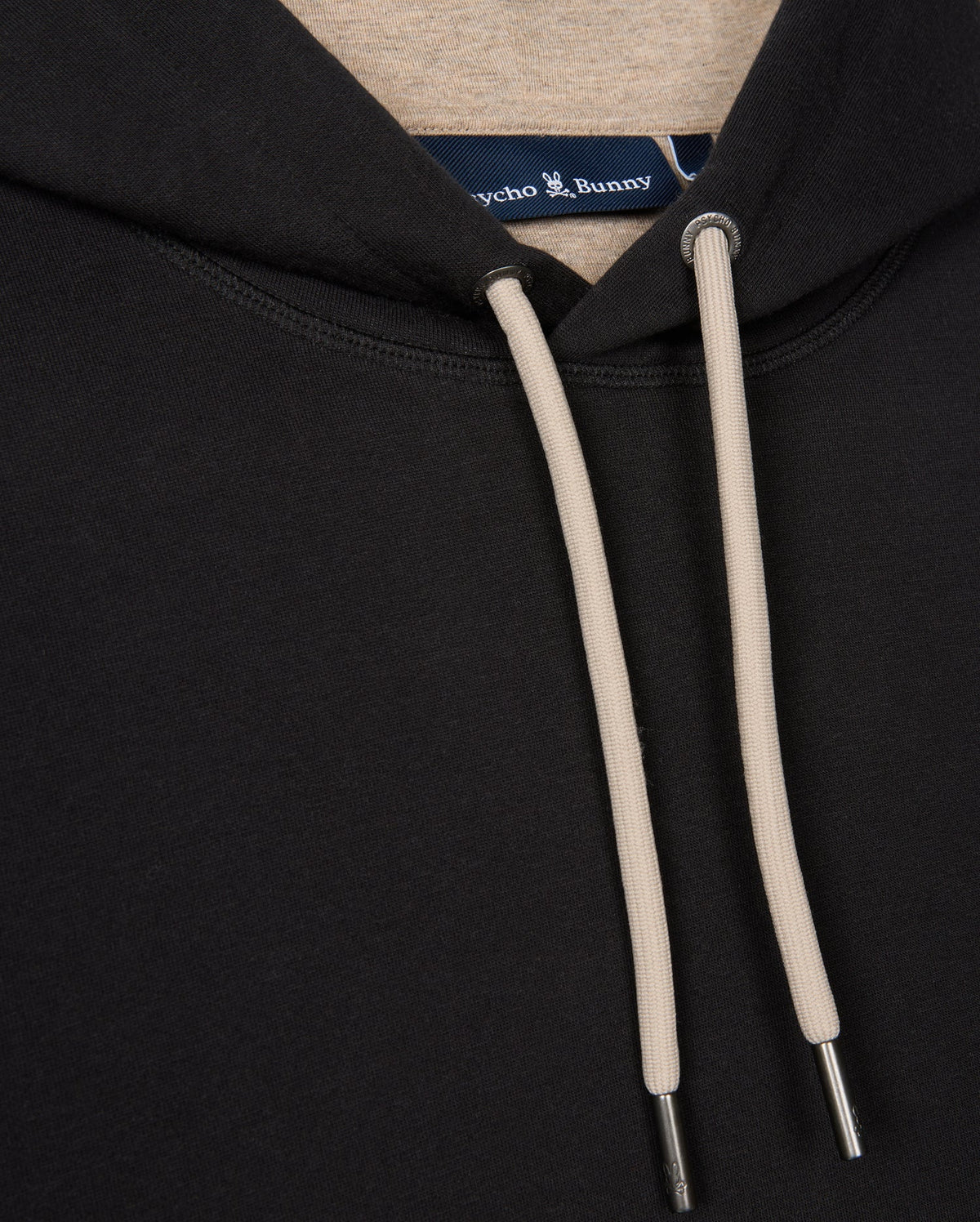Close-up of the MENS JAMES PREMIUM HOODIE, model B6H789D200, showcasing a black design with beige drawstrings and metal tips. The label sewn inside the collar reads "Psycho Bunny," accompanied by an embroidered bunny logo. This essential fleece fabric is soft and comfortable, providing both style and premium quality.
