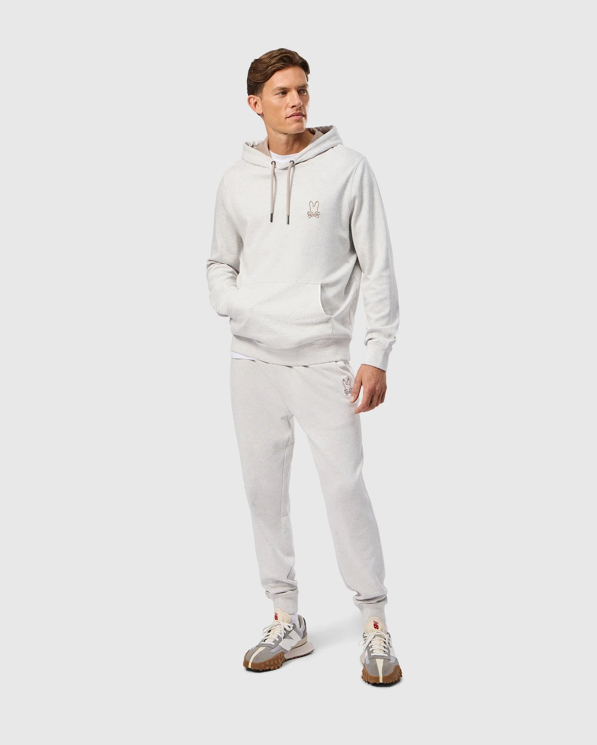 A person wearing a Psycho Bunny MENS JAMES PREMIUM HOODIE - B6H789D200 in light gray, along with matching sweatpants, is standing against a plain background. The hoodie features an embroidered bunny on the chest. The ensemble is complemented by gray and white sneakers, one hand resting casually in a pocket as the individual exudes a relaxed demeanor.