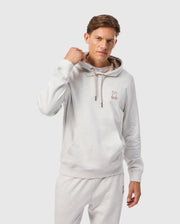 A person is wearing the MENS JAMES PREMIUM HOODIE - B6H789D200 by Psycho Bunny, featuring a light gray color and an embroidered bunny logo on the chest, with the hood strings slightly pulled. They are also sporting matching light gray essential fleece sweatpants against a plain white background.