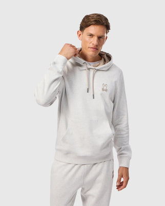 A person is wearing the MENS JAMES PREMIUM HOODIE - B6H789D200 by Psycho Bunny, featuring a light gray color and an embroidered bunny logo on the chest, with the hood strings slightly pulled. They are also sporting matching light gray essential fleece sweatpants against a plain white background.