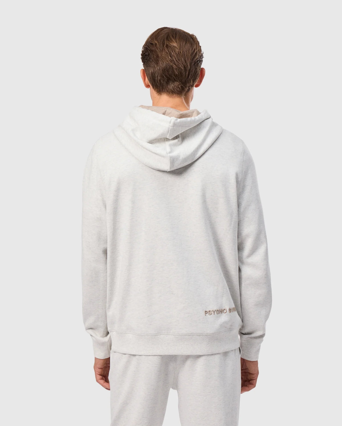 A person is seen from the back wearing a light gray MENS JAMES PREMIUM HOODIE - B6H789D200 and matching sweatpants. The hoodie prominently displays "Psycho Bunny" near the bottom, along with an embroidered bunny detail. The background is plain gray.