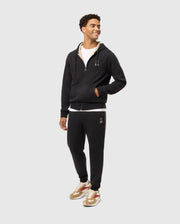 A man stands wearing a Psycho Bunny MENS JAMES PREMIUM ZIP HOODIE (B6H793D200) and matching black sweatpants, both adorned with small white logo designs. He has his left hand in his pants pocket and is smiling, looking to his right. Embracing stylish warmth, he also sports white sneakers with beige and red accents.
