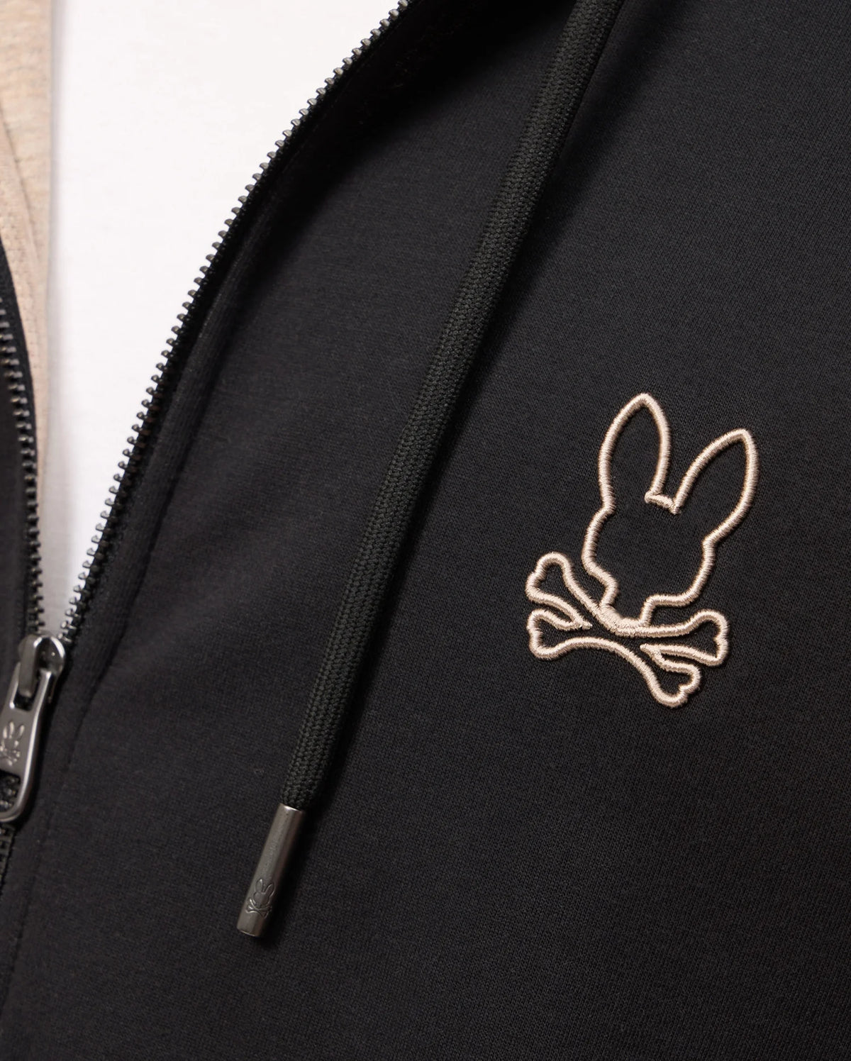 Close-up of a Psycho Bunny MENS JAMES PREMIUM ZIP HOODIE - B6H793D200 in black, featuring a white outline embroidered logo of a rabbit's head with crossed bones on the left chest. Made from premium jersey, the hoodie offers stylish warmth with visible zipper pull and drawstrings.