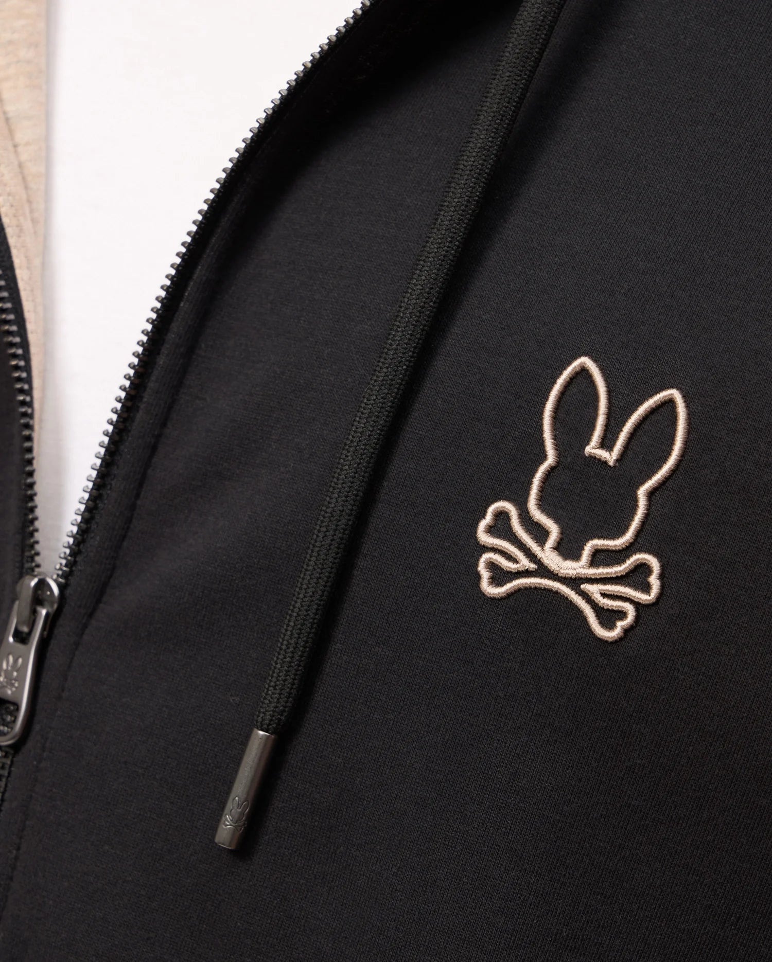 Close-up of a Psycho Bunny MENS JAMES PREMIUM ZIP HOODIE - B6H793D200 in black, featuring a white outline embroidered logo of a rabbit's head with crossed bones on the left chest. Made from premium jersey, the hoodie offers stylish warmth with visible zipper pull and drawstrings.