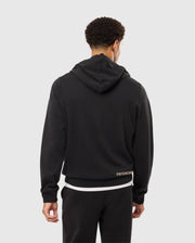 A person with short curly hair is standing with their back to the camera, wearing a black Men's James Premium Zip Hoodie (B6H793D200) from Psycho Bunny and black pants. A white T-shirt is visible underneath the stylish warmth of the hoodie, which has the word "PSYCHO" partially visible at the bottom.