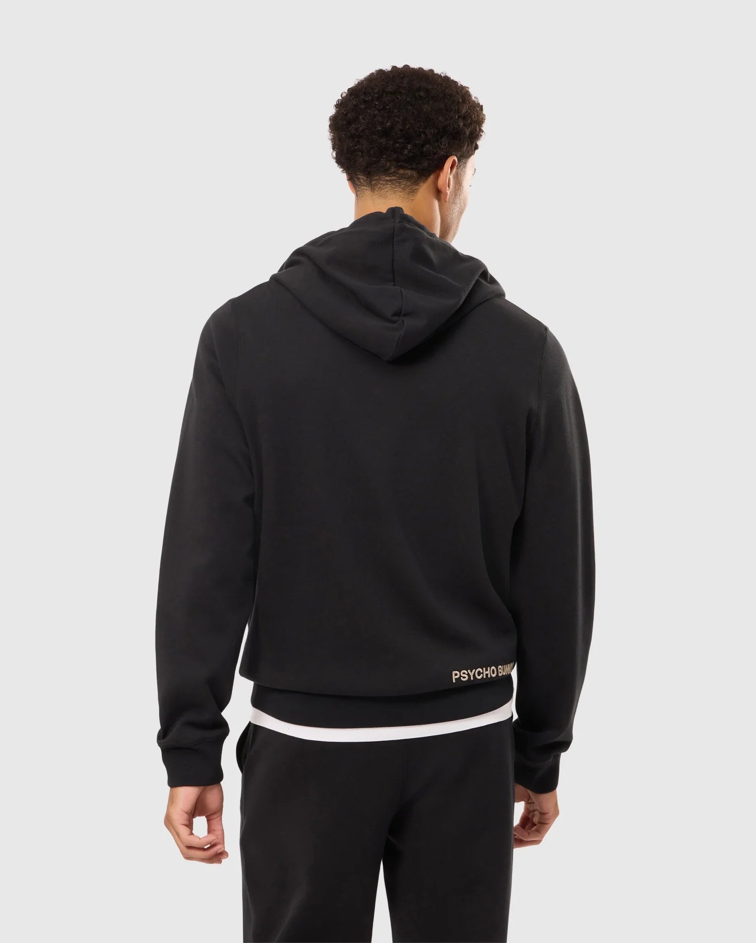 A person with short curly hair is standing with their back to the camera, wearing a black Men's James Premium Zip Hoodie (B6H793D200) from Psycho Bunny and black pants. A white T-shirt is visible underneath the stylish warmth of the hoodie, which has the word "PSYCHO" partially visible at the bottom.