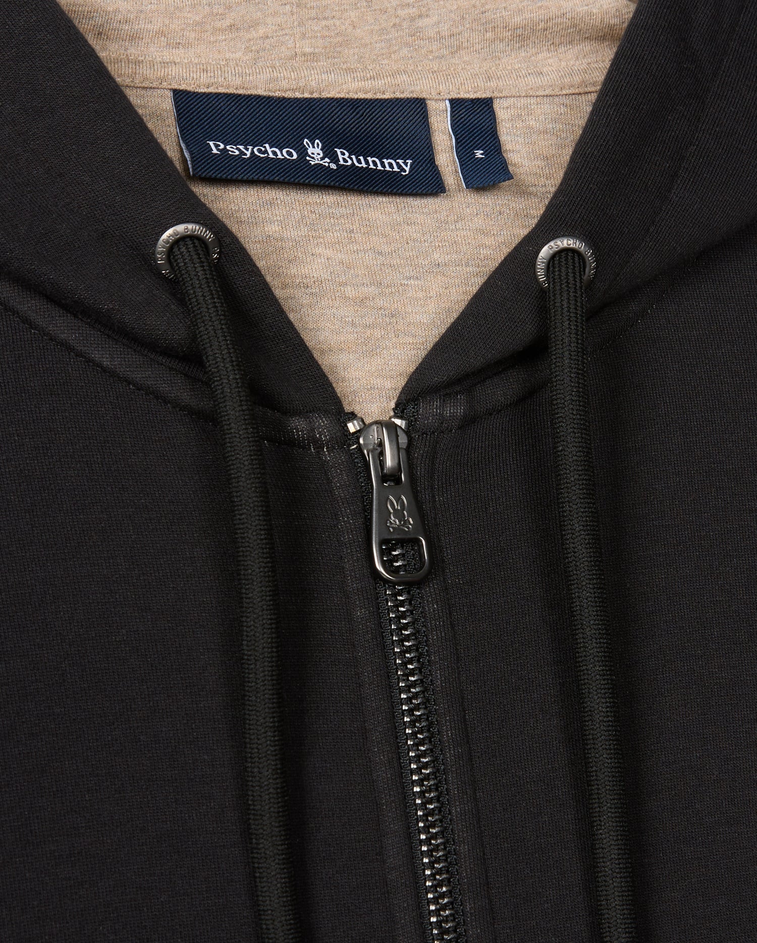 Close-up of the black MENS JAMES PREMIUM ZIP HOODIE - B6H793D200 showcasing a metal zipper, black drawstrings with metal caps, and a label inside the neckline with the brand name "Psycho Bunny" and a small bunny logo. The inner lining is a light tan color, offering stylish warmth in premium jersey fabric.