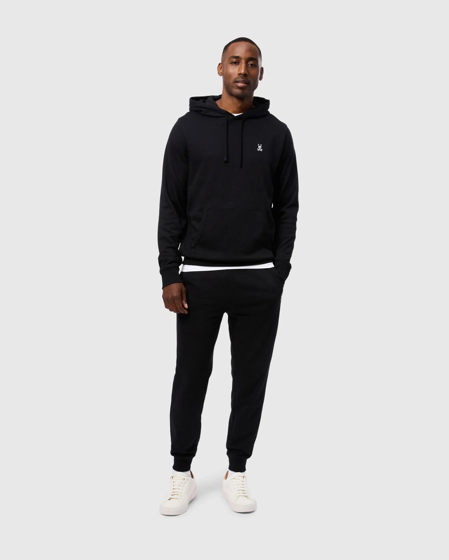 A man is standing against a plain, light grey background, wearing a Psycho Bunny MENS LEVY FLEECE HOODIE - B6H859D200 with a small emblem on the chest, black jogger pants, and white sneakers. His hands are in his hoodie pockets, and he is looking at the camera with a neutral expression.