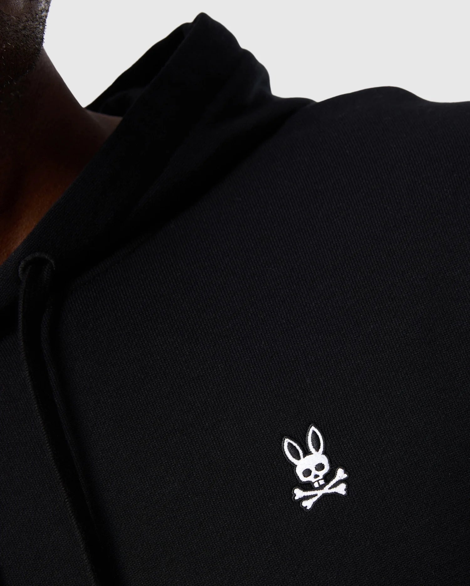 Close-up of a person wearing a Psycho Bunny Men's Levy Fleece Hoodie (B6H859D200) in black French terry, adorned with a white embroidered logo on the chest. The logo showcases a cartoon bunny with crossbones below it. The 100% cotton hoodie is slightly wrinkled, revealing only part of the person's torso and shoulder.