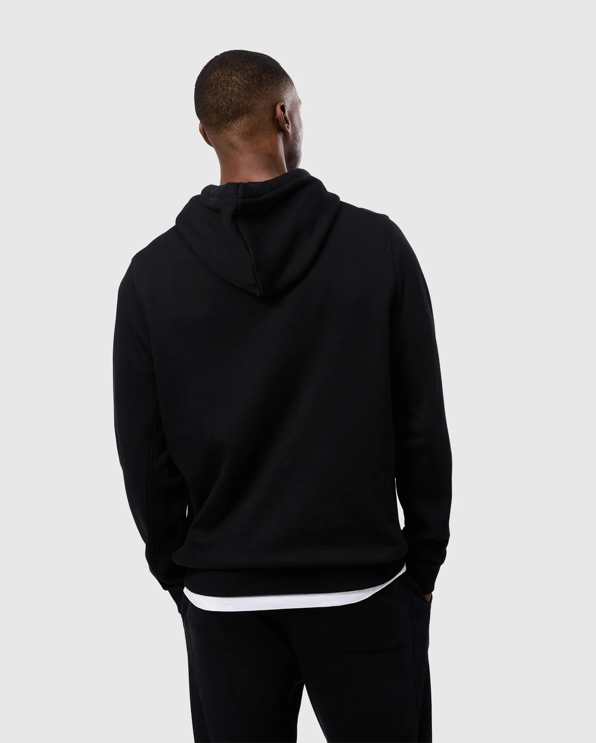 A person stands with their back to the camera, wearing a Psycho Bunny MENS LEVY FLEECE HOODIE - B6H859D200 and black pants. The individual appears to be looking to their left. The background is plain and light gray.