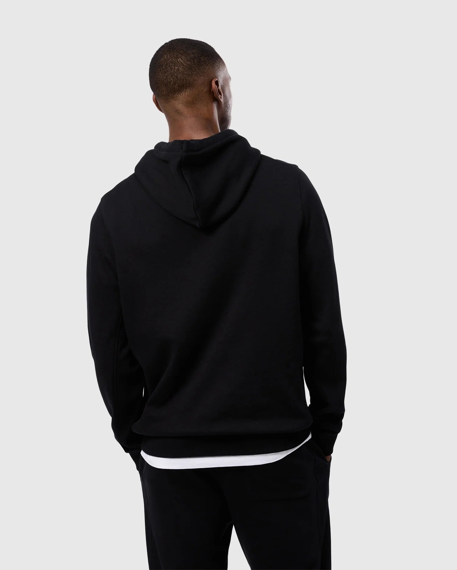 A person stands with their back to the camera, wearing a Psycho Bunny MENS LEVY FLEECE HOODIE - B6H859D200 and black pants. The individual appears to be looking to their left. The background is plain and light gray.