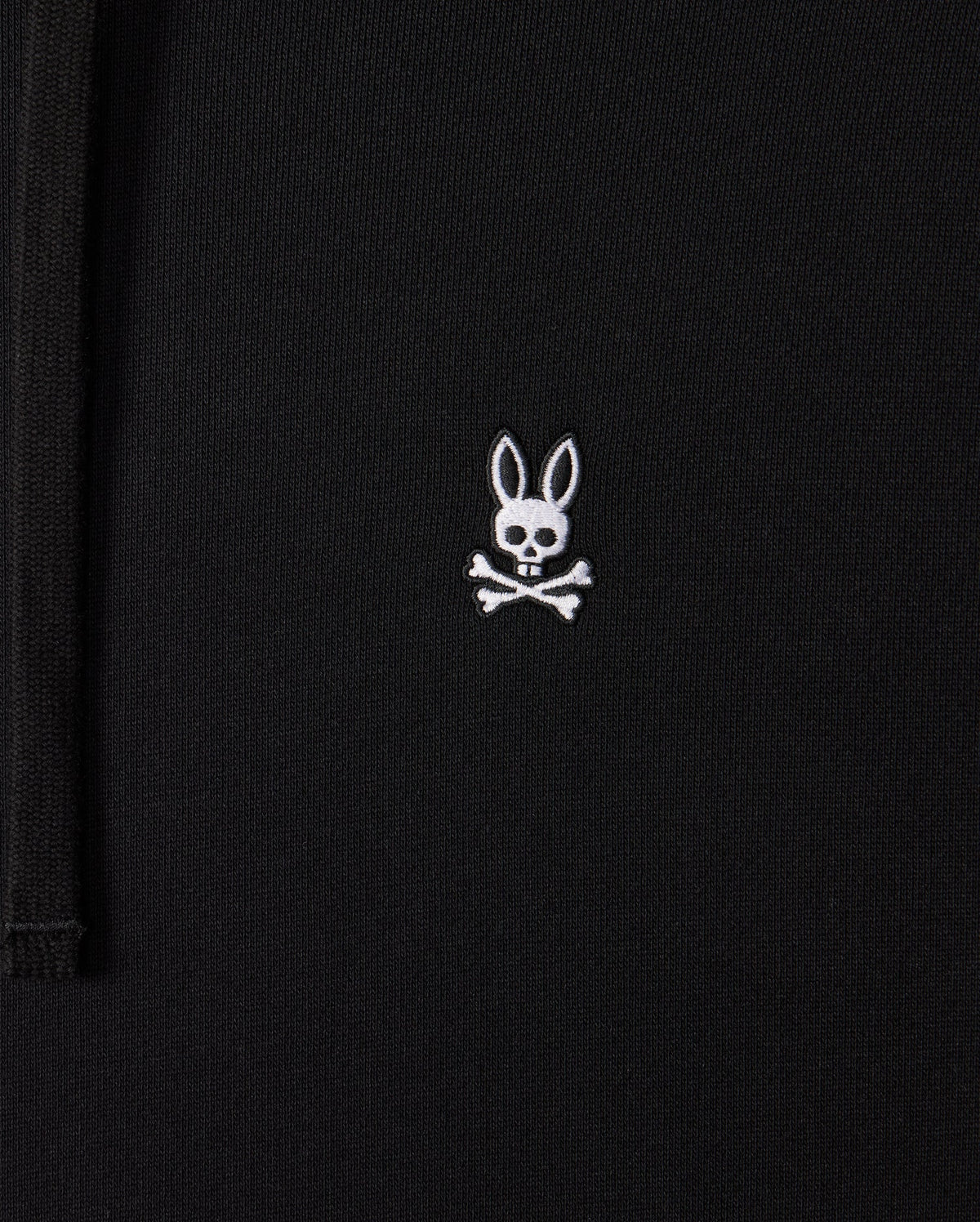 Close-up of a black Mens Levy Fleece Hoodie (B6H859D200) by Psycho Bunny, featuring a small embroidered design of a white bunny head with long ears above two crossed bones, resembling a pirate skull and crossbones. This 100% cotton garment combines comfort with unique style.