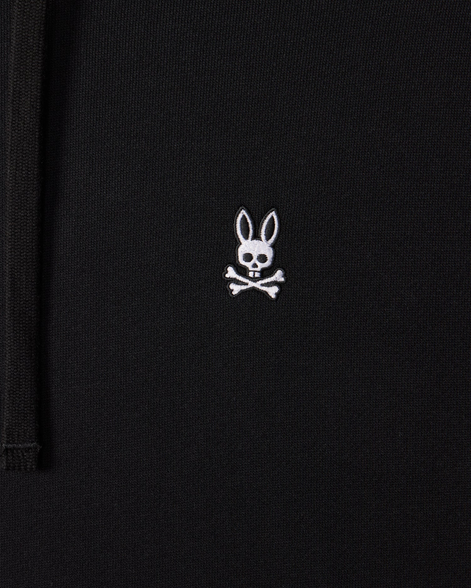 Close-up of a black Mens Levy Fleece Hoodie (B6H859D200) by Psycho Bunny, featuring a small embroidered design of a white bunny head with long ears above two crossed bones, resembling a pirate skull and crossbones. This 100% cotton garment combines comfort with unique style.