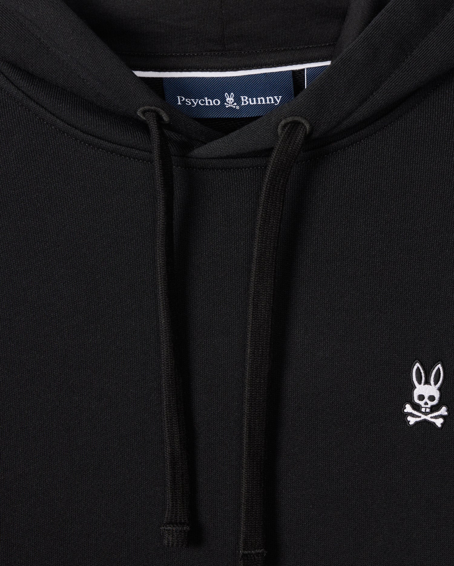 A close-up of the MENS LEVY FLEECE HOODIE - B6H859D200 from Psycho Bunny, featuring a small white embroidered logo of a bunny skull and crossbones on the left chest, showcases its sleek black French terry fabric. The 100% cotton label inside reads "Psycho Bunny," and drawstrings are visible in the foreground, making it an essential piece.