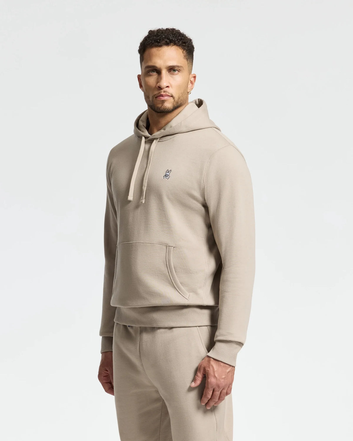 A man wearing a Psycho Bunny MENS LEVY ESSENTIAL FLEECE HOODIE crafted from soft, 100% cotton fleece, along with matching sweatpants, stands against a plain background. The beige hoodie highlights a small embroidered design on the chest. He is facing forward with his hands by his sides.
