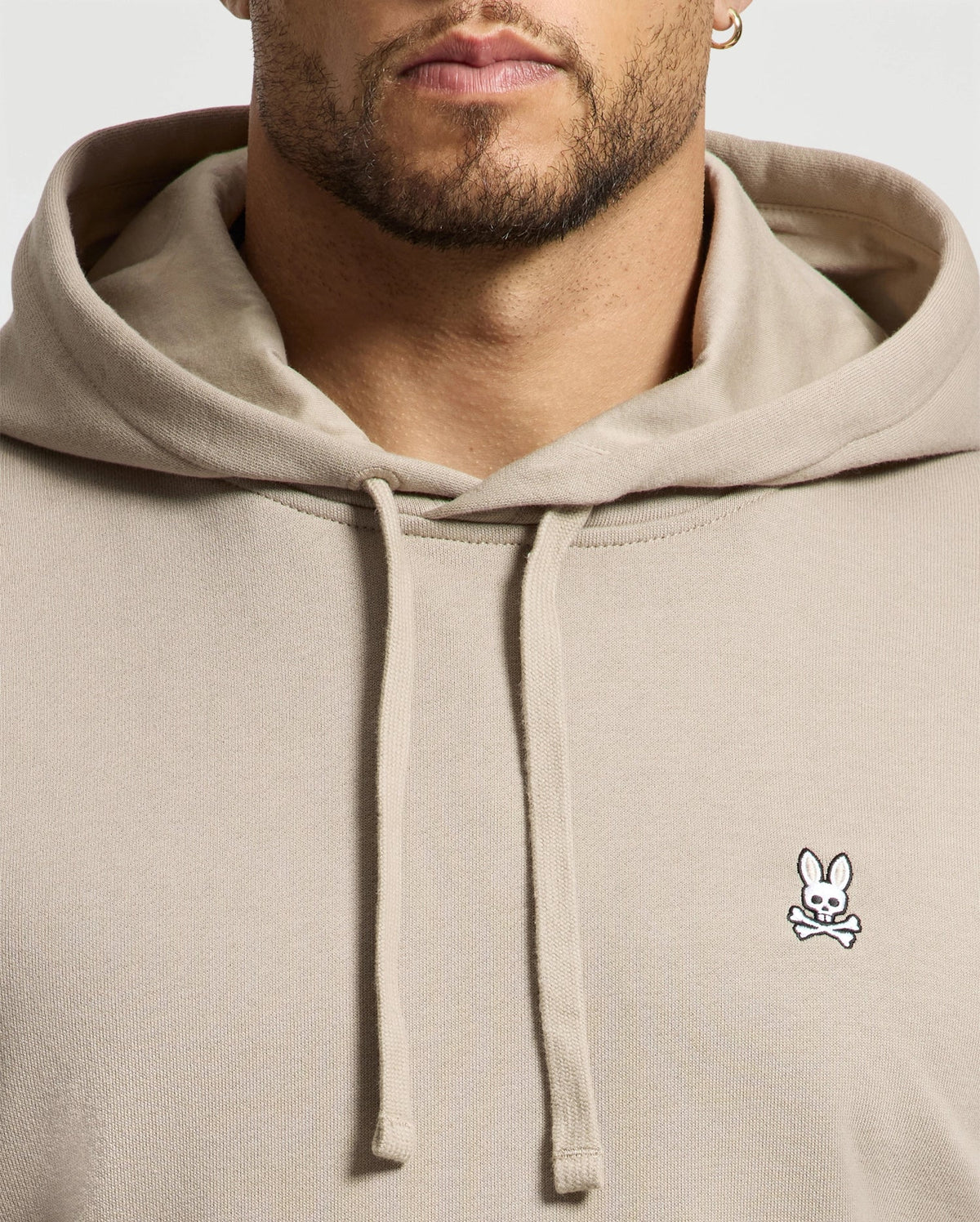 The individual is dressed in a beige Men's Levy Essential Fleece Hoodie (B6H859D200) from Psycho Bunny, made from premium 100% cotton fleece and featuring a small embroidered logo of a bunny with crossed bones on the chest, highlighting its distinguished design details and high-quality fabric.