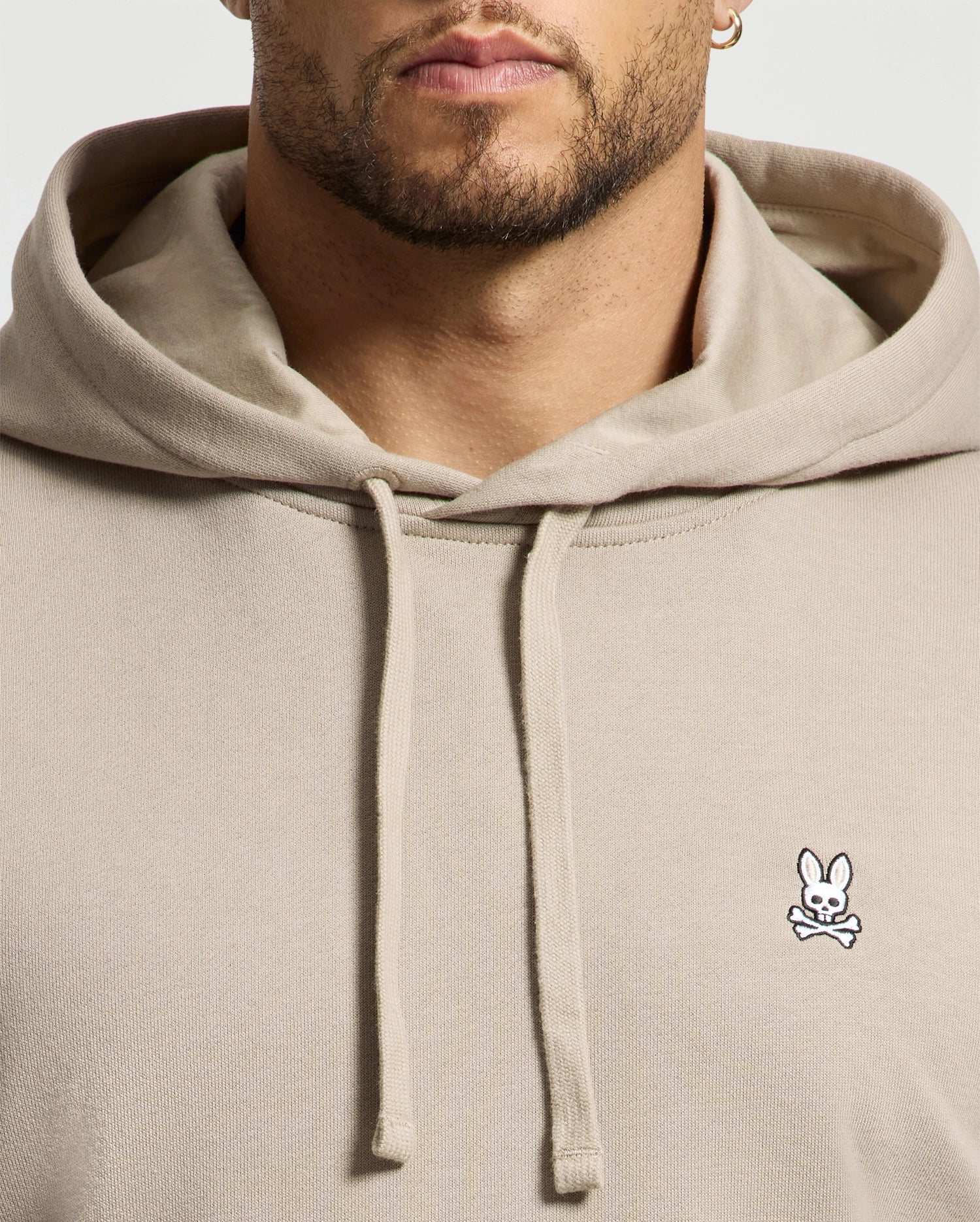 The individual is dressed in a beige Men's Levy Essential Fleece Hoodie (B6H859D200) from Psycho Bunny, made from premium 100% cotton fleece and featuring a small embroidered logo of a bunny with crossed bones on the chest, highlighting its distinguished design details and high-quality fabric.