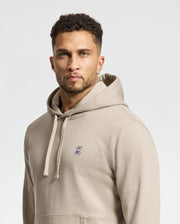 A person with short curly hair wearing a beige Psycho Bunny MENS LEVY ESSENTIAL FLEECE HOODIE - B6H859D200, crafted from men's 100% cotton, stands against a light background, looking directly at the camera.