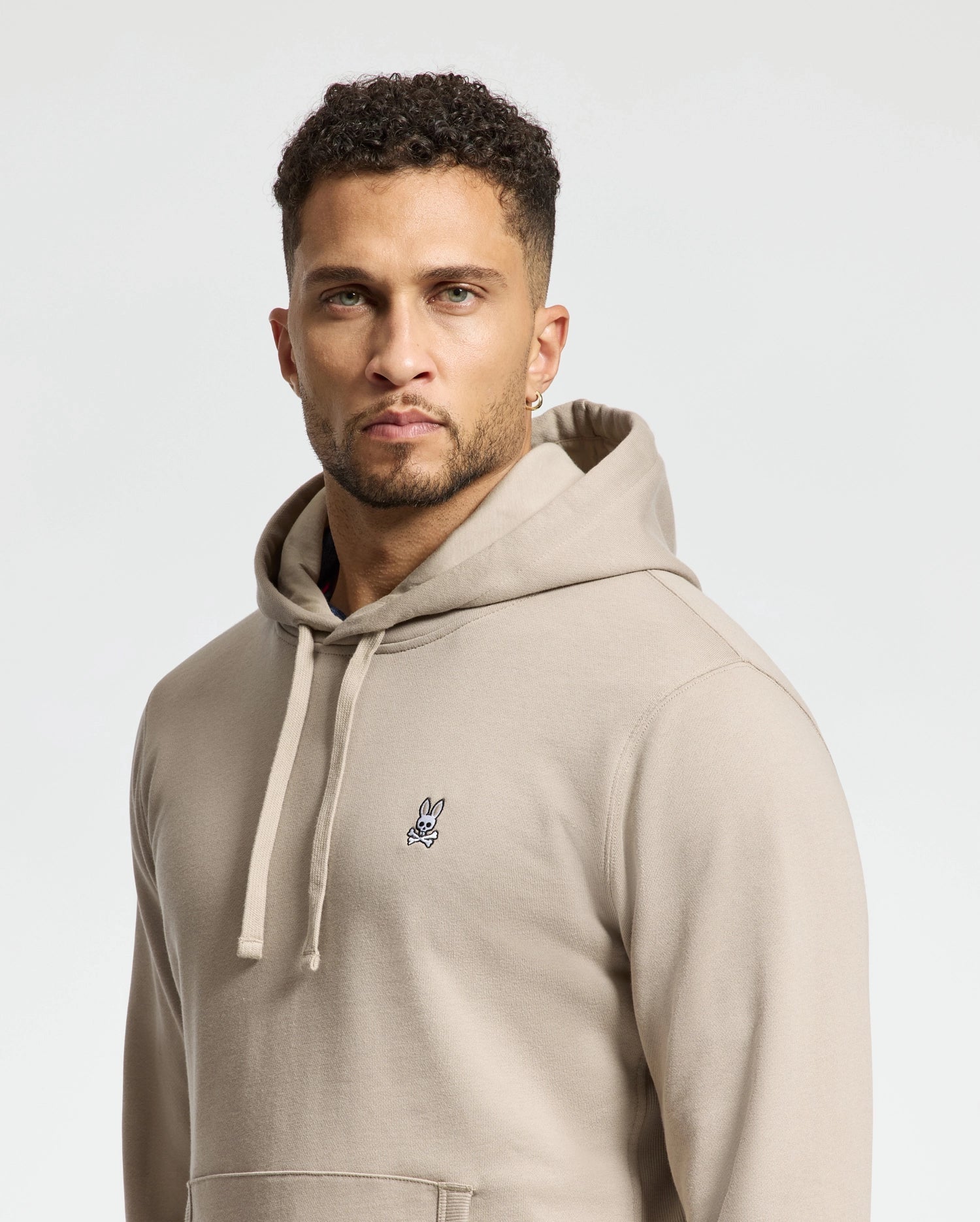 A person with short curly hair wearing a beige Psycho Bunny MENS LEVY ESSENTIAL FLEECE HOODIE - B6H859D200, crafted from men's 100% cotton, stands against a light background, looking directly at the camera.