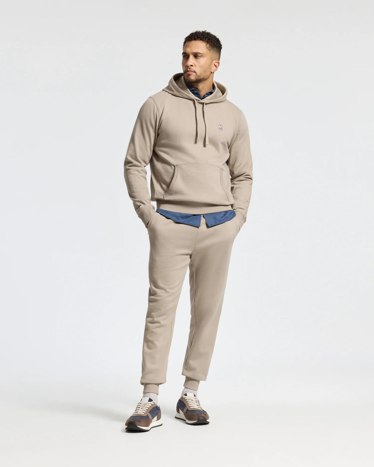 A man casually stands with his hands in his pockets, sporting a Psycho Bunny Men's Levy Essential Fleece Hoodie (100% cotton) and matching sweatpants over a denim shirt. He's wearing sneakers while looking slightly to the side against a plain background, adding a touch of comfort to his laid-back style.