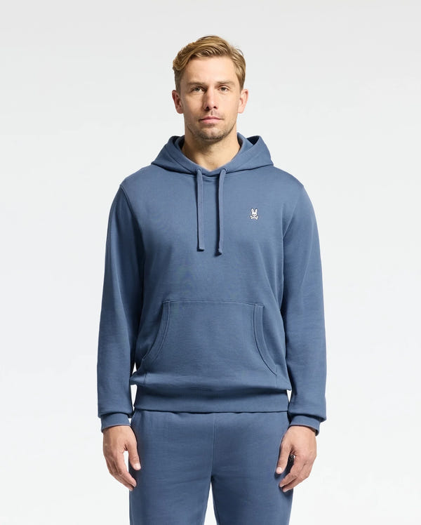 A man in a Psycho Bunny MENS LEVY ESSENTIAL FLEECE HOODIE and matching sweatpants stands against a white backdrop. The 100% cotton hoodie boasts an embroidered chest logo, with his hands relaxed at his sides, showcasing effortless style.
