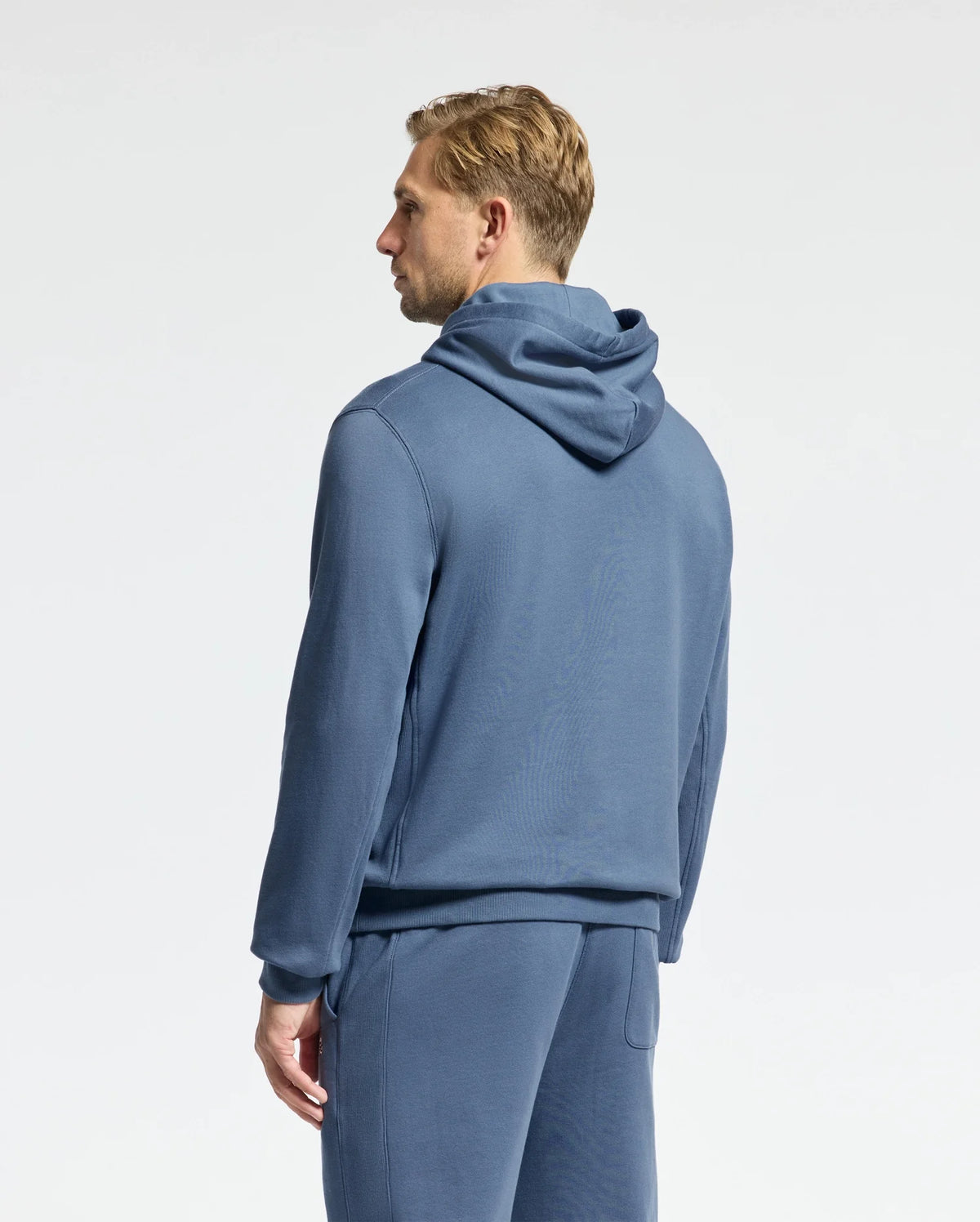 A person with short blond hair faces away from the camera, wearing Psycho Bunny's Men's Levy Essential Fleece Hoodie and matching pants in soft blue. Crafted from 100% cotton, the ensemble is set against a plain, light-colored background.