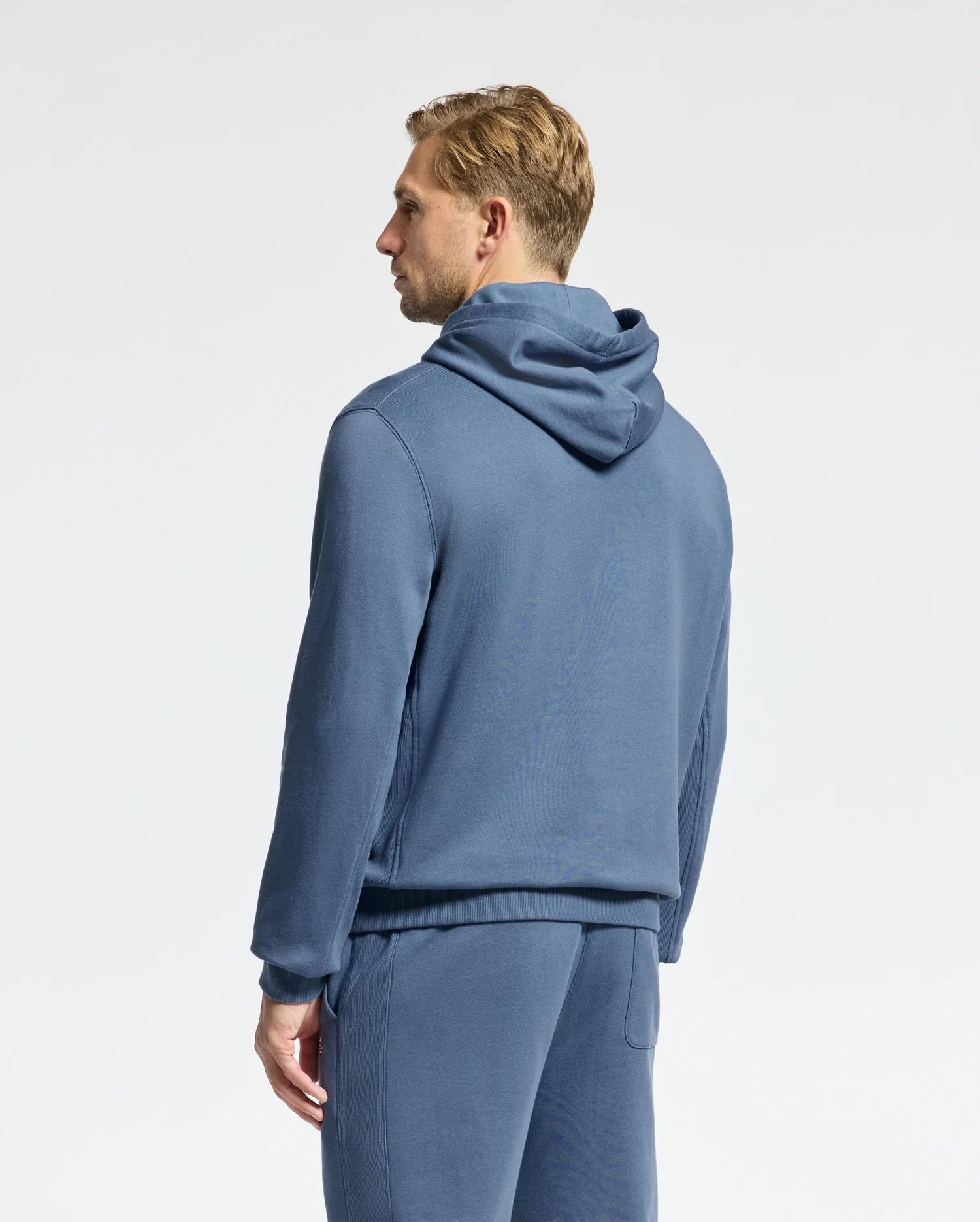 A person with short blond hair faces away from the camera, wearing Psycho Bunny's Men's Levy Essential Fleece Hoodie and matching pants in soft blue. Crafted from 100% cotton, the ensemble is set against a plain, light-colored background.