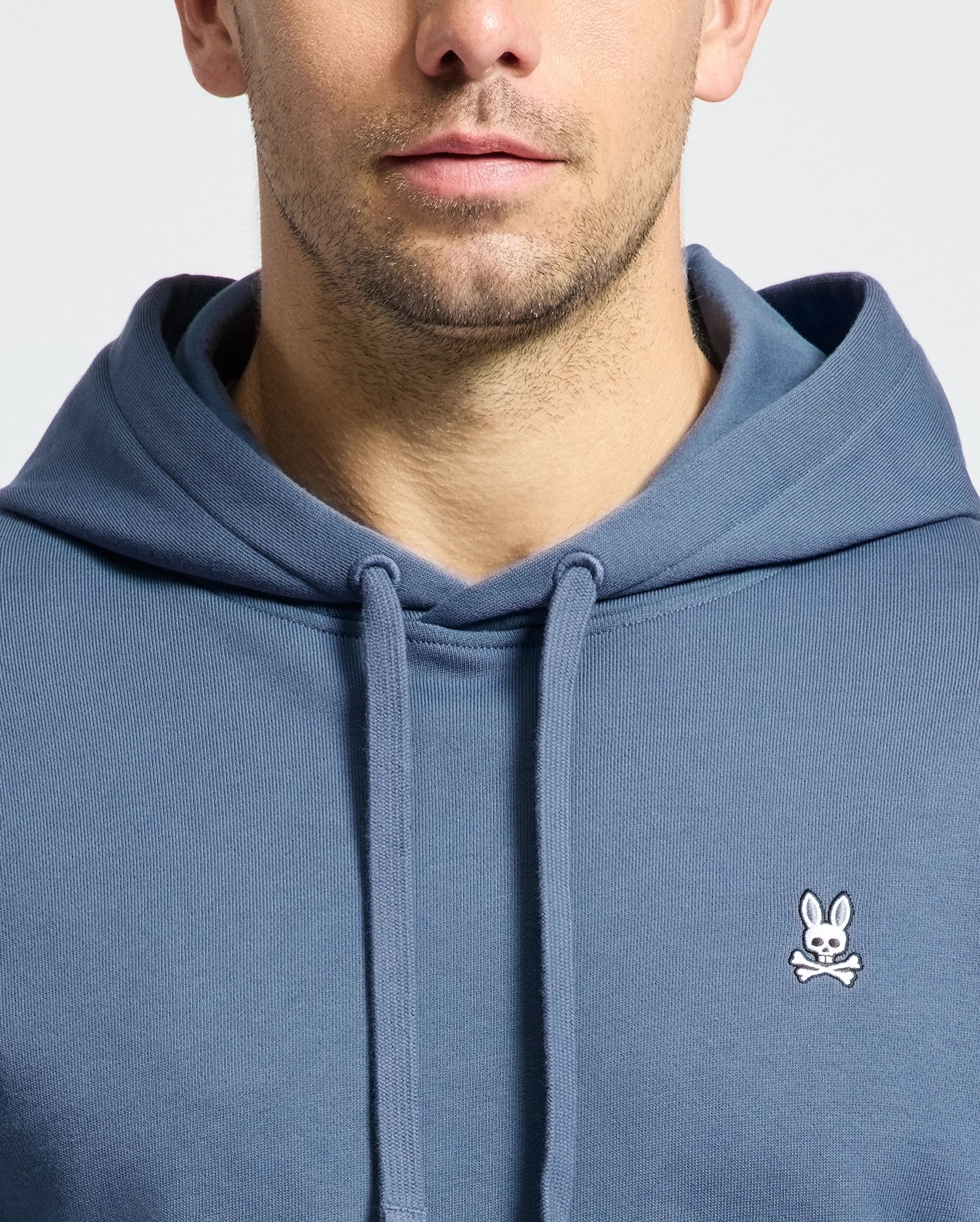 A person in a Psycho Bunny MENS LEVY ESSENTIAL FLEECE HOODIE with a bunny emblem on the chest. The cropped shot highlights men's fashion, showing only the lower face and torso. Made of 100% cotton, this blue hoodie features drawstrings and a smooth texture.