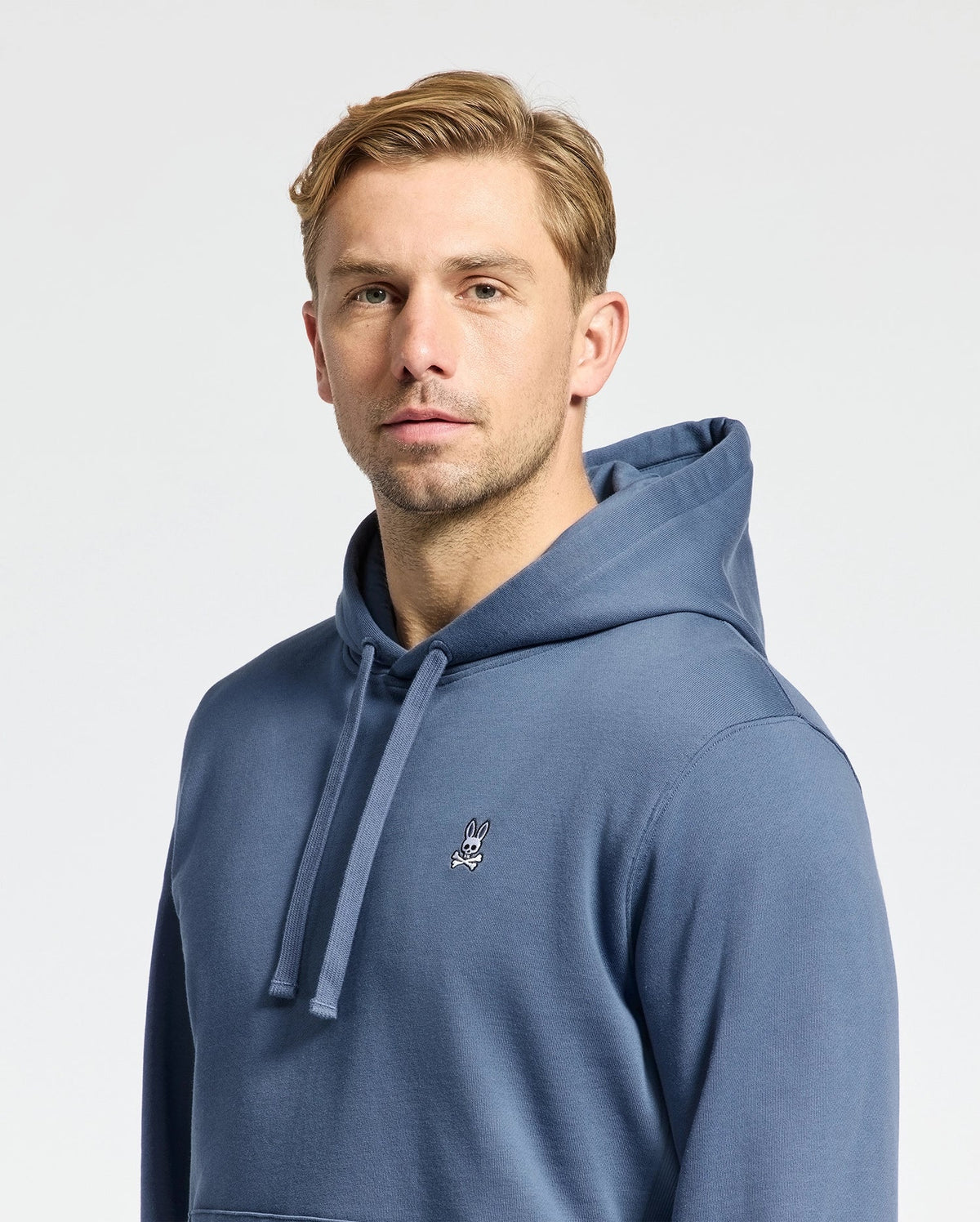 A short-haired blond person wears a Psycho Bunny Men's Levy Essential Fleece Hoodie (B6H859D200) in blue, with a small embroidered rabbit logo on the chest. Made from 100% cotton, this hoodie is a men's fashion staple. The background is plain white.