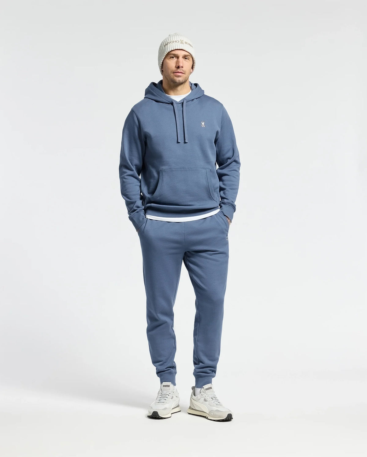 A person dressed in a Psycho Bunny MENS LEVY ESSENTIAL FLEECE HOODIE, joggers, white sneakers, and a white knit beanie stands with hands in pockets against a plain background, embodying men's fashion.