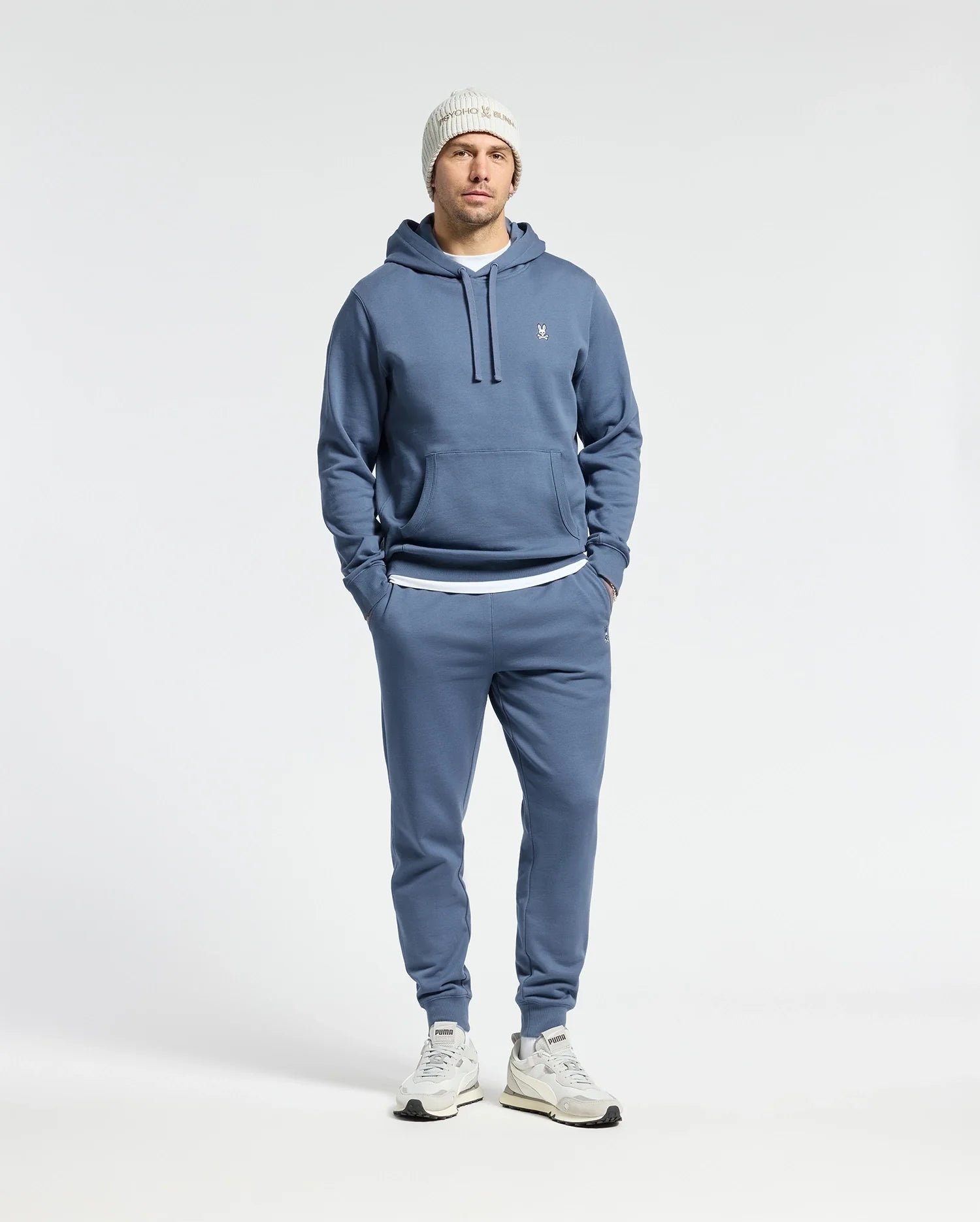 A person dressed in a Psycho Bunny MENS LEVY ESSENTIAL FLEECE HOODIE, joggers, white sneakers, and a white knit beanie stands with hands in pockets against a plain background, embodying men's fashion.