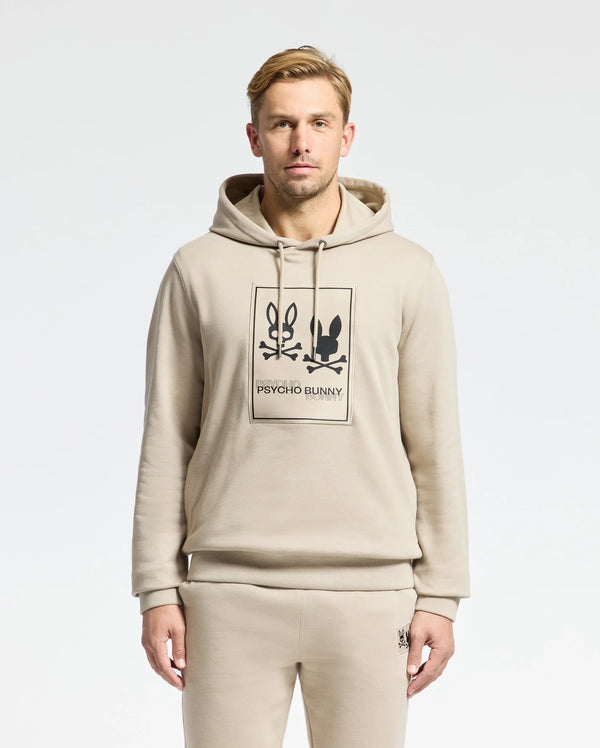 A man wears a beige Psycho Bunny MENS MONROE TWILL PATCH FLEECE HOODIE (B6H926D200) and matching sweatpants, featuring two bunny faces and "PSYCHO BUNNY" text. He stands facing forward against a plain light background.