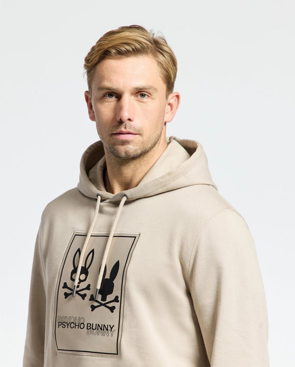 A man with short blond hair wears the Psycho Bunny MENS MONROE TWILL PATCH FLEECE HOODIE, featuring a black graphic of two bunny skulls and crossbones with "PSYCHO BUNNY" text, looking directly at the camera with a neutral expression.