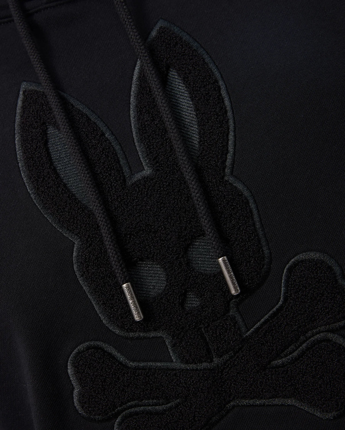 Close-up of a black Psycho Bunny MENS PACIFIC CHENILLE HOODIE - B6H930D200 featuring an embroidered design of a bunny skull with crossbones beneath it. The hoodie strings have metallic tips. The design is tonal, with the embroidery just slightly raised and darker than the 100% cotton fabric.