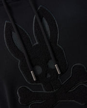 Close-up of a black Psycho Bunny MENS PACIFIC CHENILLE HOODIE - B6H930D200 featuring an embroidered design of a bunny skull with crossbones beneath it. The hoodie strings have metallic tips. The design is tonal, with the embroidery just slightly raised and darker than the 100% cotton fabric.