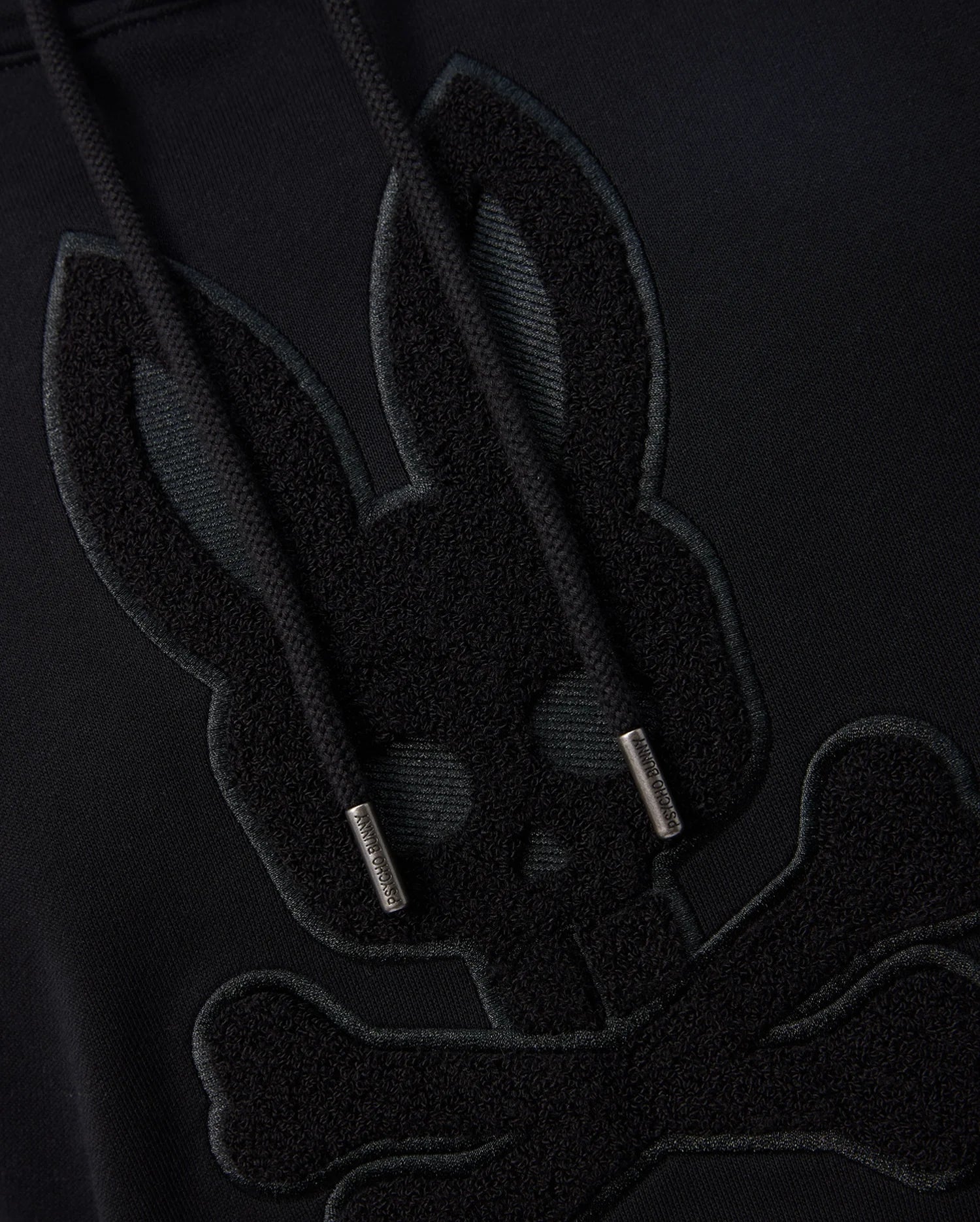 Close-up of a black Psycho Bunny MENS PACIFIC CHENILLE HOODIE - B6H930D200 featuring an embroidered design of a bunny skull with crossbones beneath it. The hoodie strings have metallic tips. The design is tonal, with the embroidery just slightly raised and darker than the 100% cotton fabric.