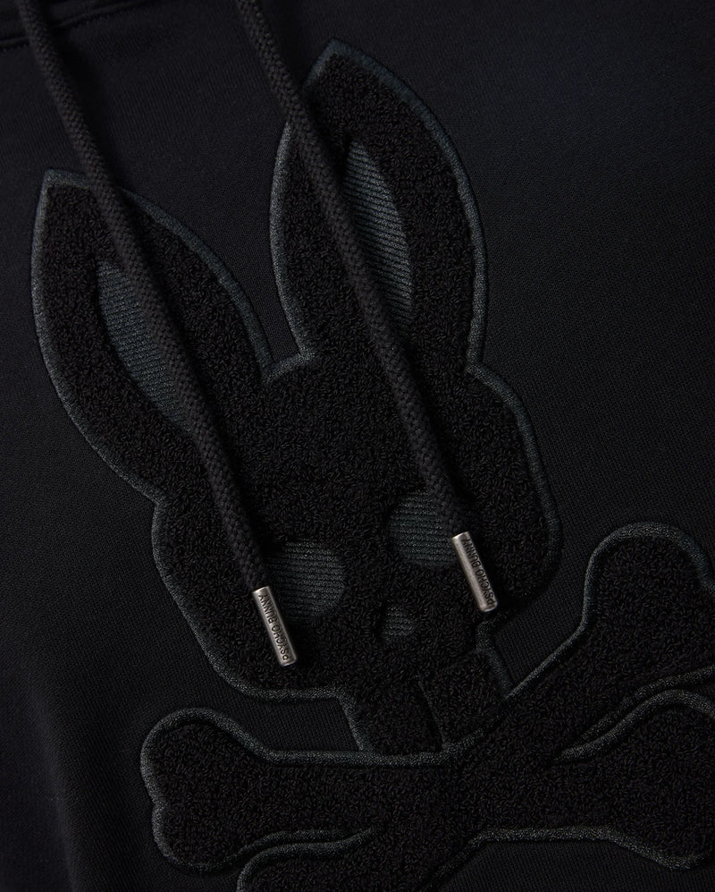 Close-up of a black Psycho Bunny MENS PACIFIC CHENILLE HOODIE - B6H930D200 featuring an embroidered design of a bunny skull with crossbones beneath it. The hoodie strings have metallic tips. The design is tonal, with the embroidery just slightly raised and darker than the 100% cotton fabric.