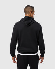 A man stands facing away from the camera, wearing a black Psycho Bunny MENS PACIFIC CHENILLE HOODIE - B6H930D200 and black pants with a white shirt peeking out from underneath. His hair is short and neatly trimmed. The background is plain and light-colored.