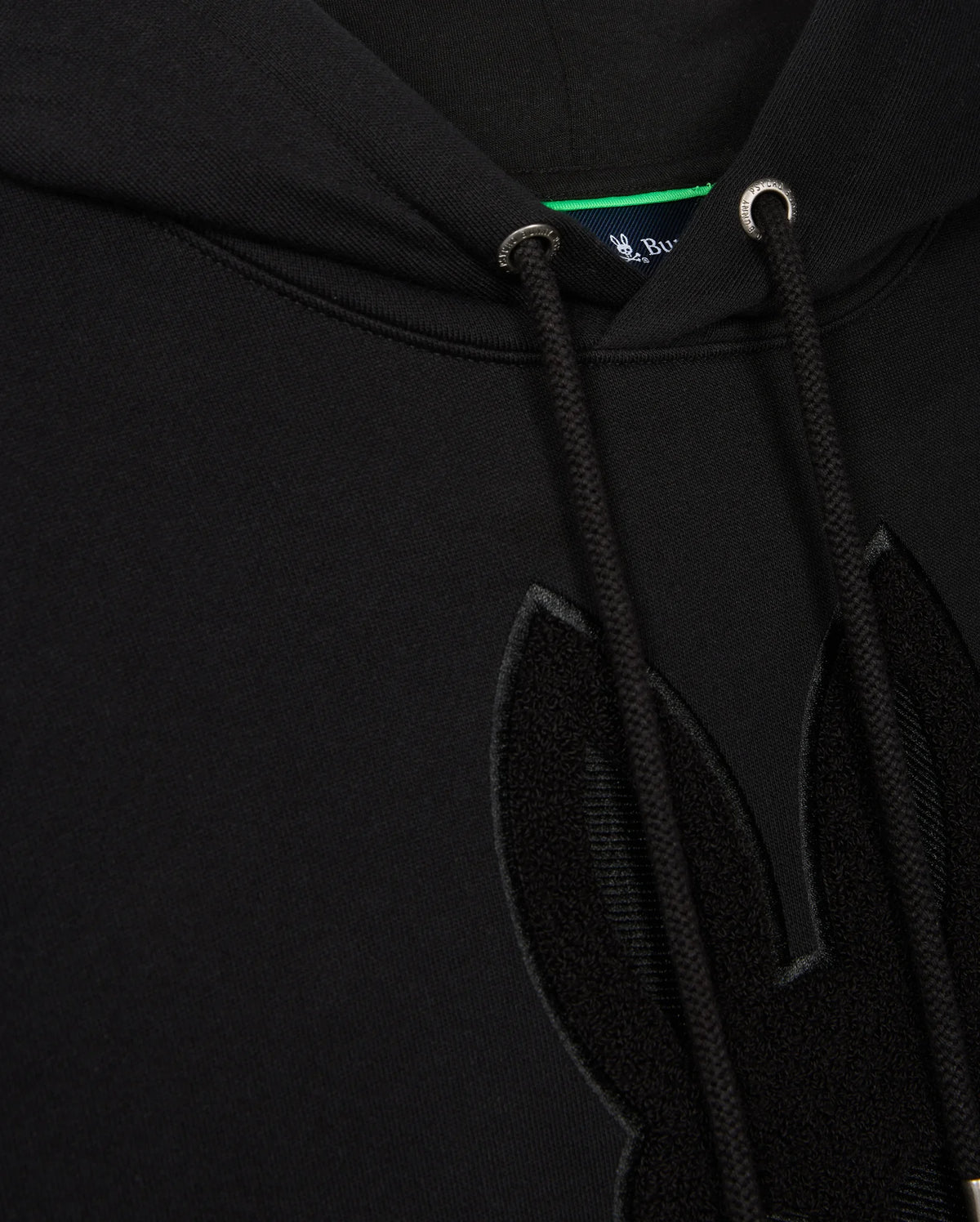 Close-up of the Psycho Bunny Men's Pacific Chenille Hoodie (B6H930D200) in black French terry, showcasing a textured chenille embroidery patch of bunny ears on the front. This 100% cotton hoodie includes drawstrings with metal eyelets, and a small portion of the blue inner tag can be seen at the top.