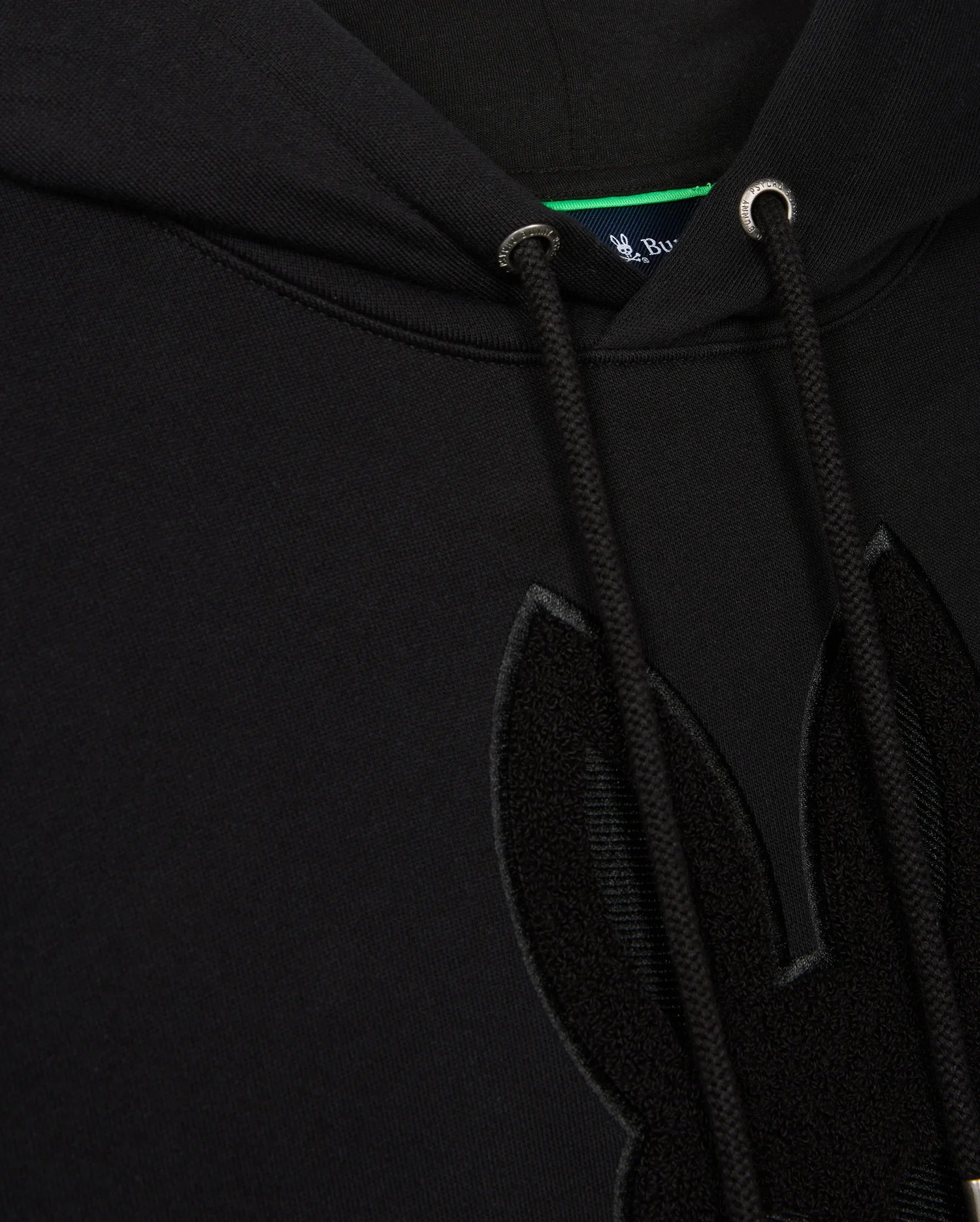 Close-up of the Psycho Bunny Men's Pacific Chenille Hoodie (B6H930D200) in black French terry, showcasing a textured chenille embroidery patch of bunny ears on the front. This 100% cotton hoodie includes drawstrings with metal eyelets, and a small portion of the blue inner tag can be seen at the top.