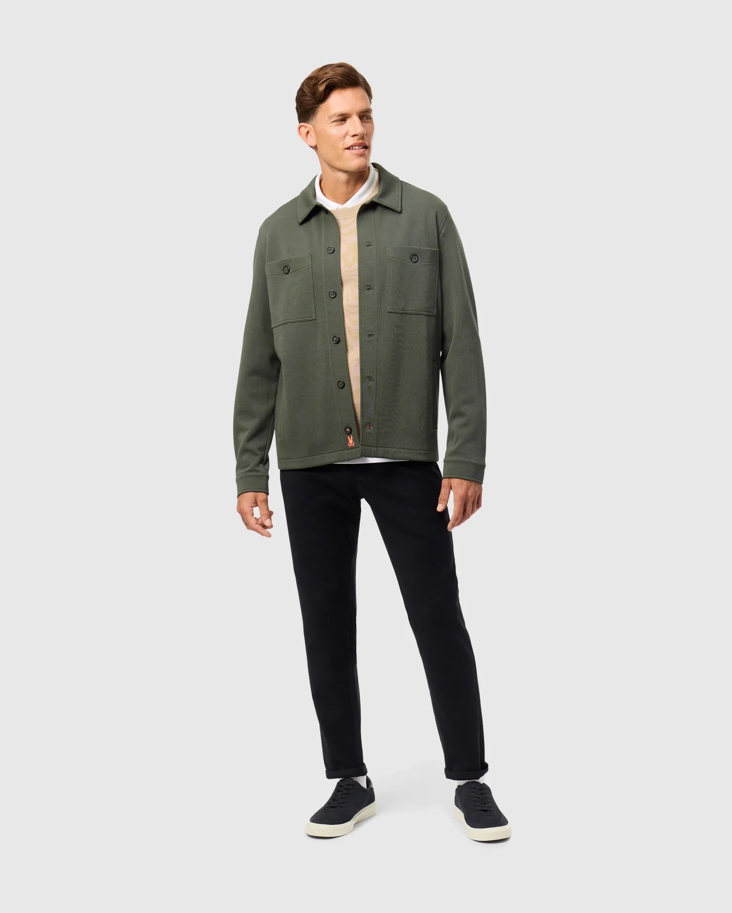 Wearing a green Men's Bedford Shirt Jacket by Psycho Bunny over a beige cotton-blend shirt, black pants, and black sneakers, the person stands against a plain background. Slightly angled with a neutral expression, they embody an understated urban vibe.