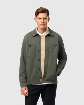 Wearing a green Psycho Bunny MENS BEDFORD SHIRT JACKET - B6J290B200 over a beige and white sweater, the person with short brown hair stands against a plain background. The cotton-blend jacket features two front pockets and a small logo near the hem, embodying an urban feel as they look at the camera.