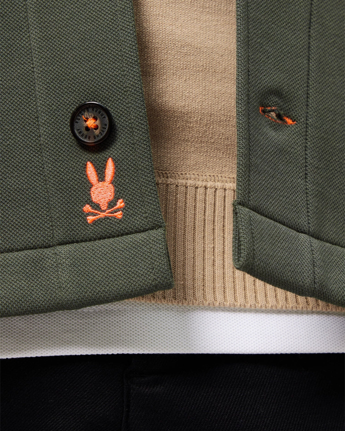 Close-up of the green Psycho Bunny Mens Bedford Shirt Jacket - B6J290B200 over a beige sweater. An embroidered orange bunny with crossed bones adds an urban vibe to the buttonhole, accompanied by a black button and a peek of black pants below.