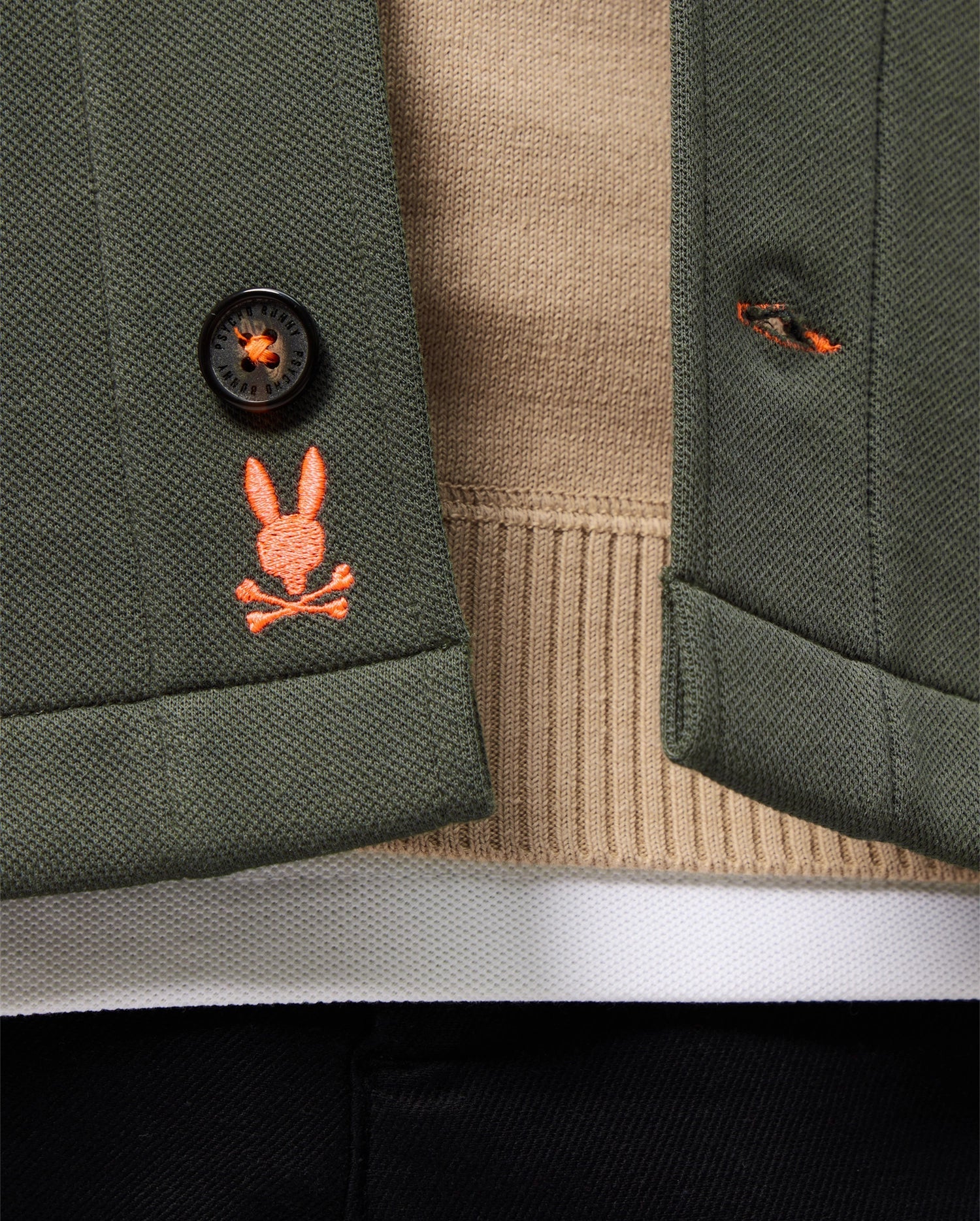 Close-up of the green Psycho Bunny Mens Bedford Shirt Jacket - B6J290B200 over a beige sweater. An embroidered orange bunny with crossed bones adds an urban vibe to the buttonhole, accompanied by a black button and a peek of black pants below.