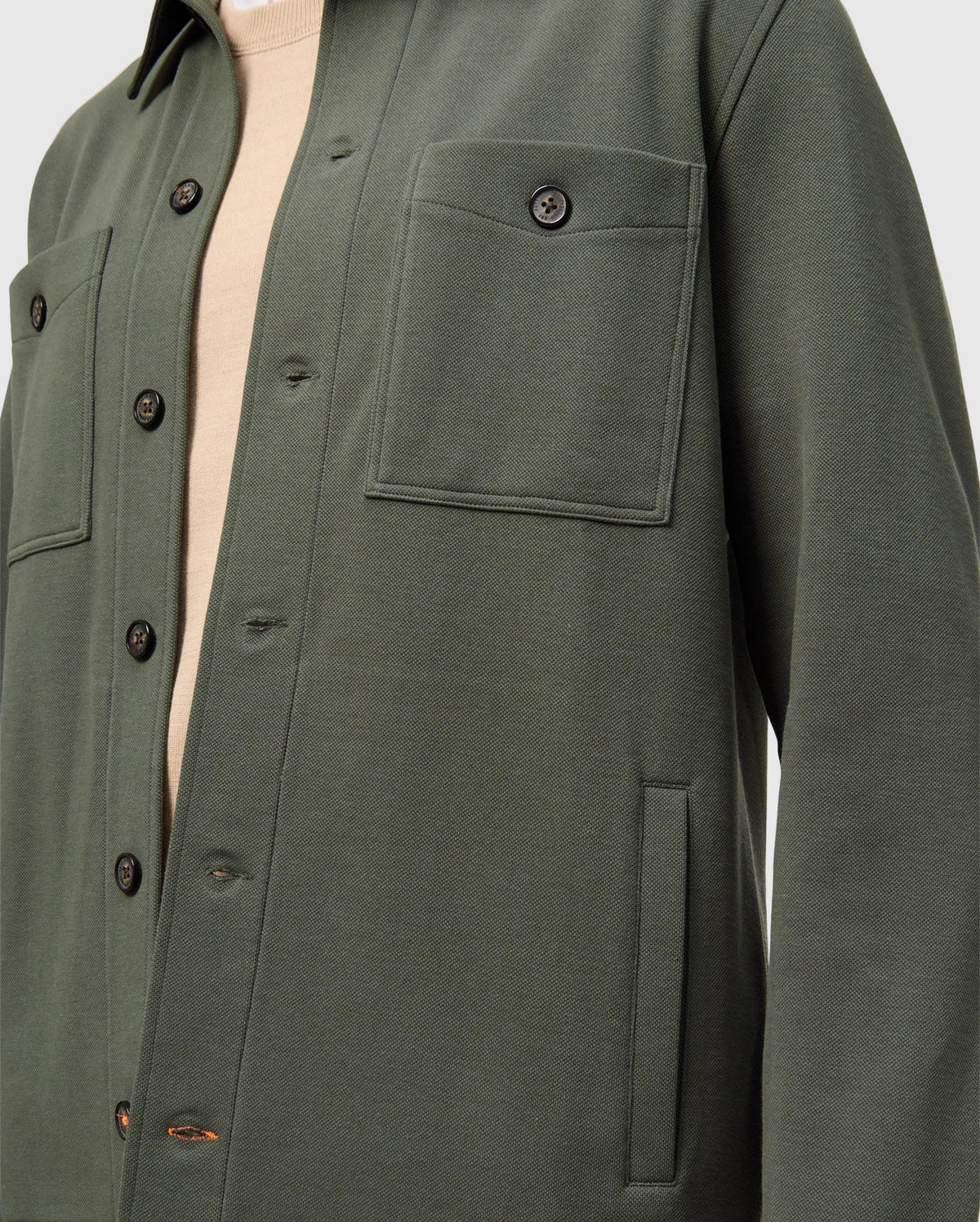 A close-up of someone wearing the Psycho Bunny MENS BEDFORD SHIRT JACKET (B6J290B200) in dark green, featuring black buttons, two front pockets, and long sleeves over a beige top. The focus is on the urban-style upper half of the jacket.