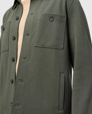 A close-up of someone wearing the Psycho Bunny MENS BEDFORD SHIRT JACKET (B6J290B200) in dark green, featuring black buttons, two front pockets, and long sleeves over a beige top. The focus is on the urban-style upper half of the jacket.