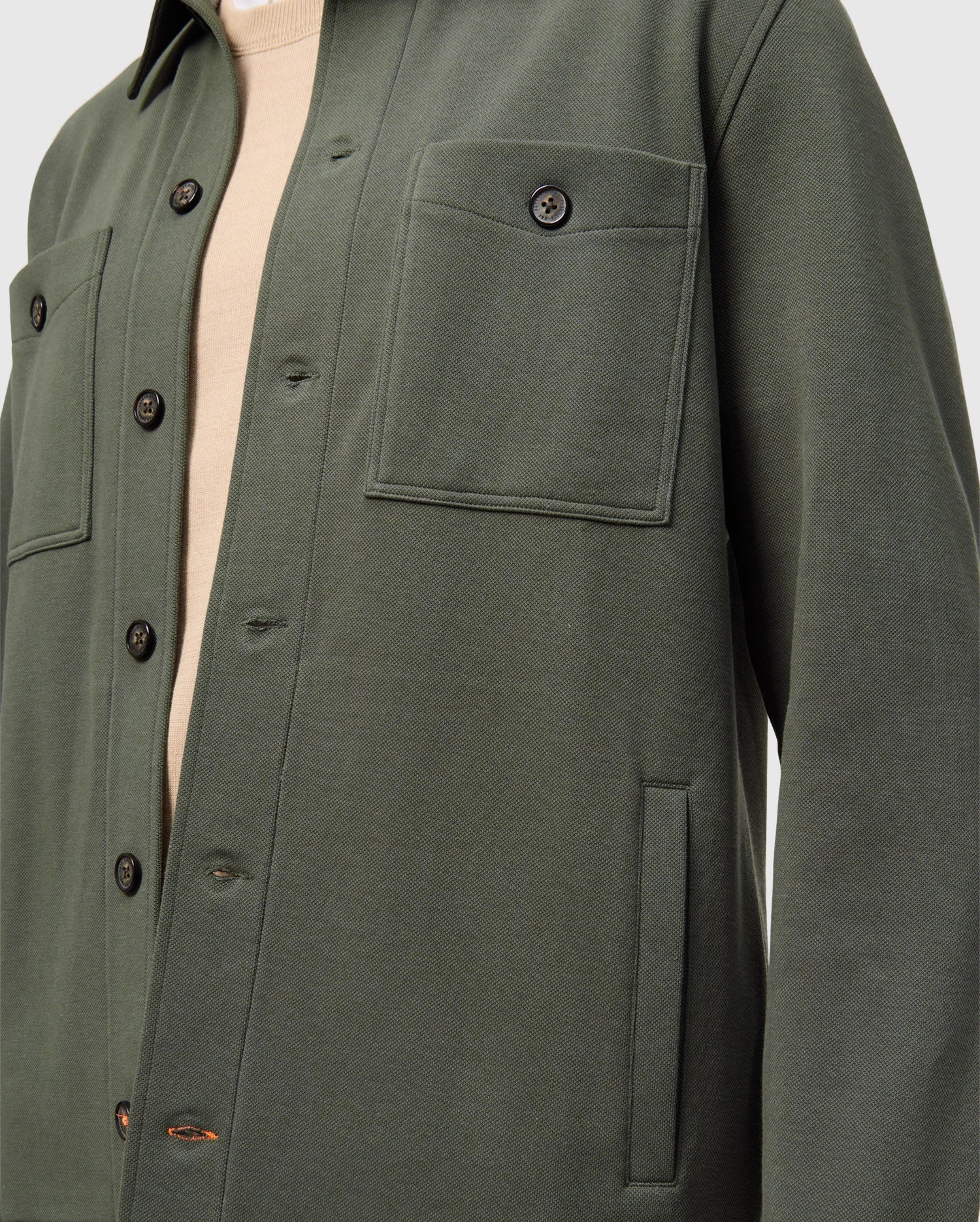 A close-up of someone wearing the Psycho Bunny MENS BEDFORD SHIRT JACKET (B6J290B200) in dark green, featuring black buttons, two front pockets, and long sleeves over a beige top. The focus is on the urban-style upper half of the jacket.