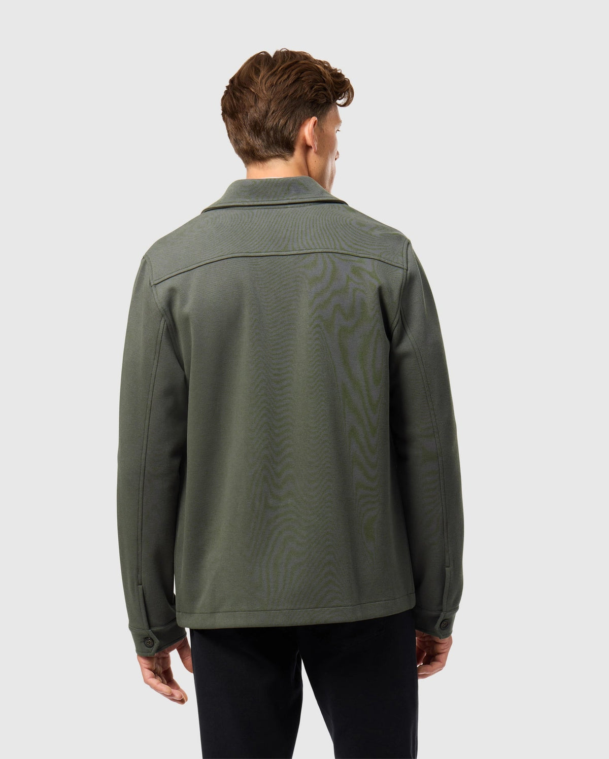 A person with short brown hair faces away from the camera, showcasing an urban vibe in Psycho Bunny's dark green cotton-blend Men's Bedford Shirt Jacket (B6J290B200) paired with black pants against a light gray backdrop.