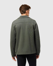 A person with short brown hair faces away from the camera, showcasing an urban vibe in Psycho Bunny's dark green cotton-blend Men's Bedford Shirt Jacket (B6J290B200) paired with black pants against a light gray backdrop.